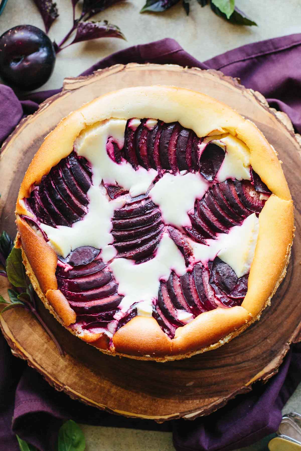 Overhead view of a whole Italian ricotta plum cheesecake on a wooden board, with fanned plum slices arranged in a starburst pattern over creamy filling.