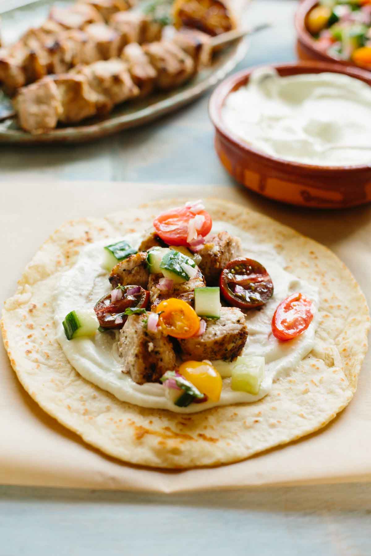 Open pita bread topped with a layer of tzatziki sauce, grilled pork cubes, cherry tomatoes, cucumber, and red onion, ready to be folded into a sandwich.