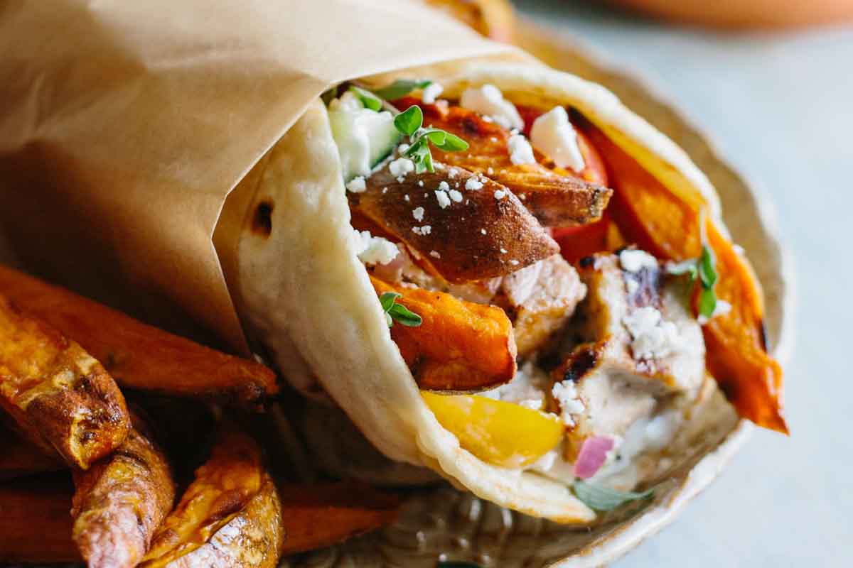 Wrapped pork souvlaki pita sandwich filled with grilled pork, sweet potato fries, cherry tomatoes, red onion, and crumbled feta, served with extra fries on the side.