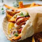 Close-up of a pork souvlaki pita wrap with sweet potato fries, pork skewers, cherry tomatoes, feta, and fresh herbs, wrapped in parchment paper on a ceramic plate.
