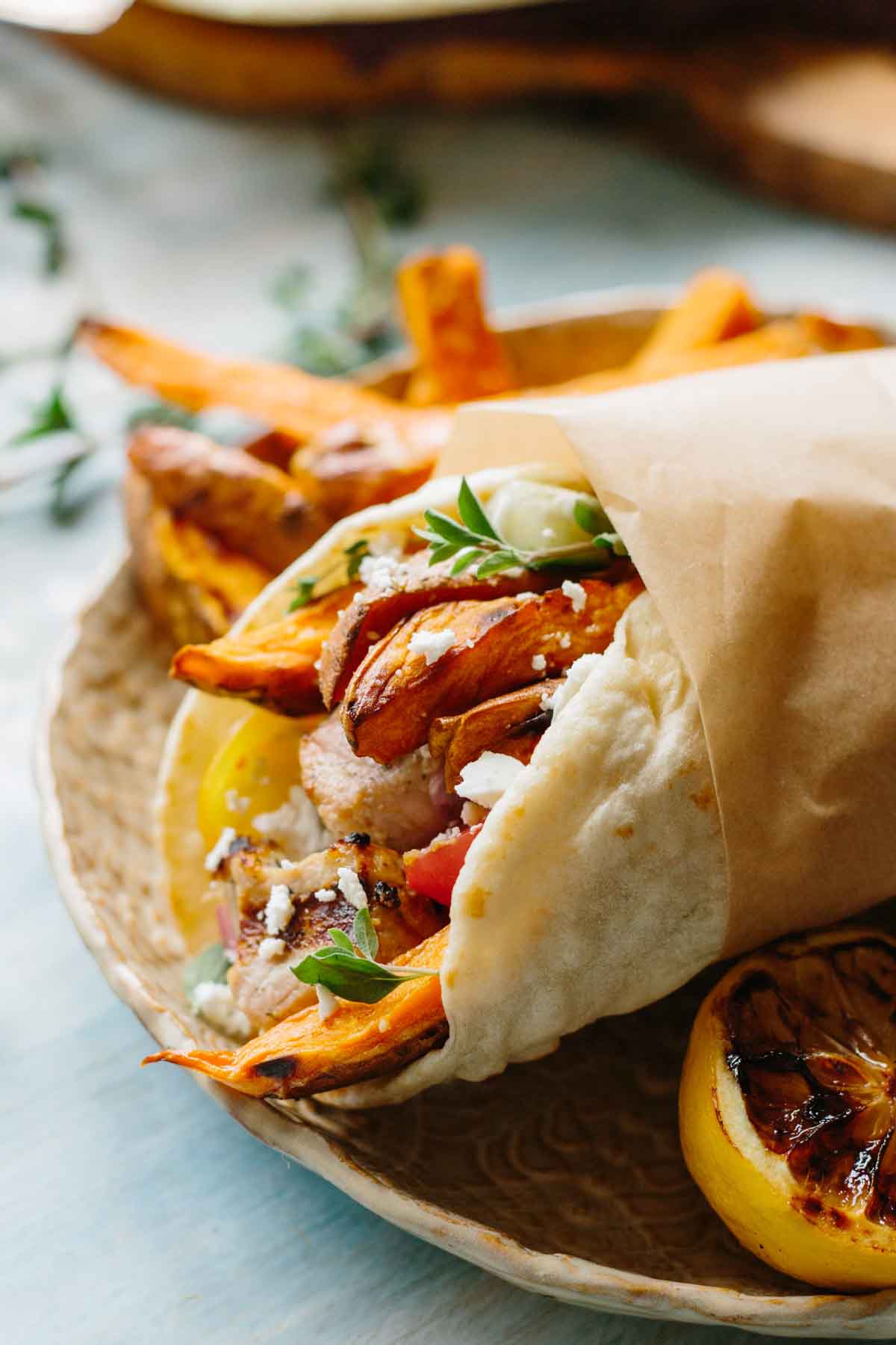 Close-up of a pork souvlaki pita wrap with sweet potato fries, pork skewers, cherry tomatoes, feta, and fresh herbs, wrapped in parchment paper on a ceramic plate.