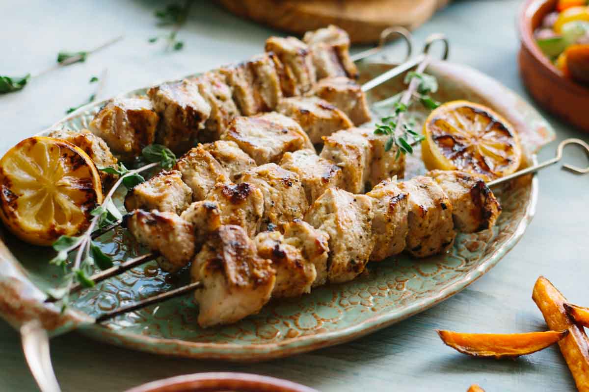Juicy grilled pork souvlaki skewers resting on a decorative platter with charred lemon halves and fresh herbs, served with salad and sweet potato fries on the side.