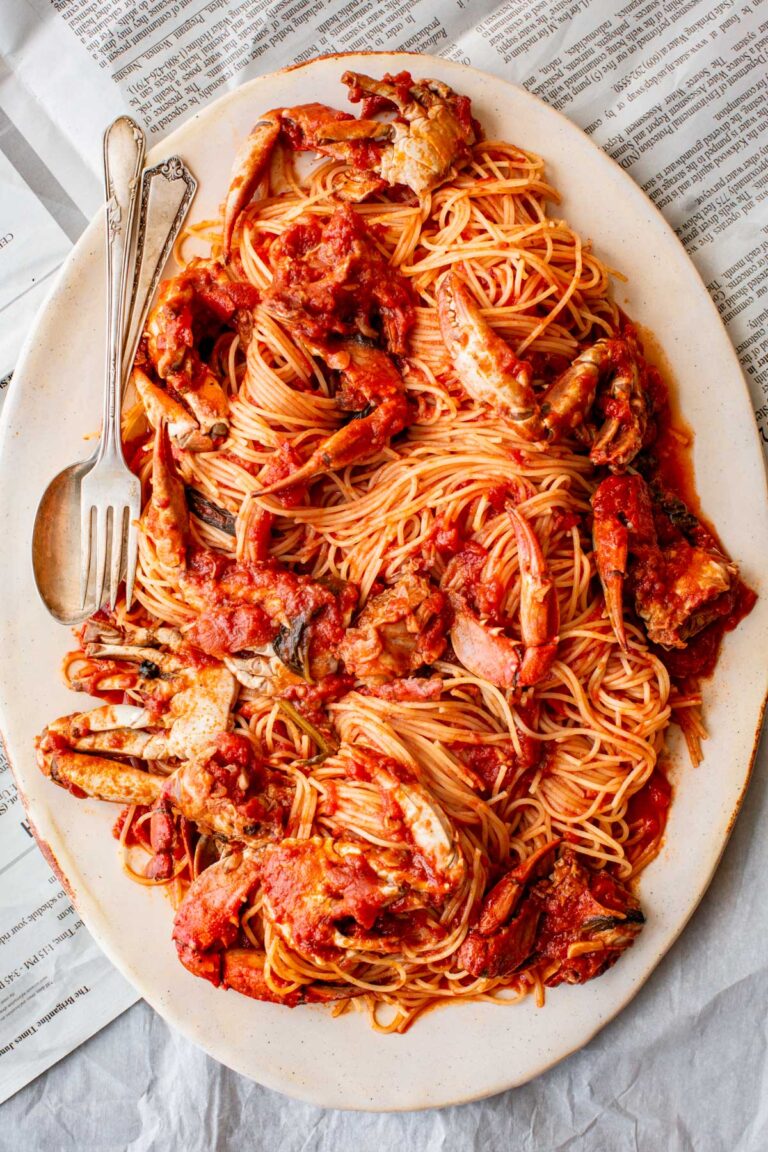 Large oval platter filled with spaghetti and crab pieces in a rich red tomato sauce, served with a fork and spoon on the side, resting on newspaper.