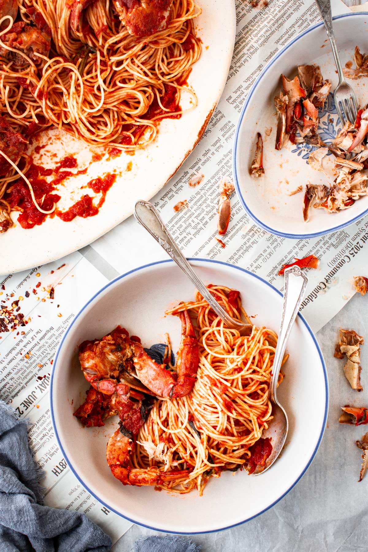 Two bowls of spaghetti with crab in tomato sauce, one partly eaten, next to the large serving platter and scattered crab shells on newspaper.