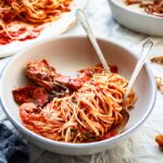 Close-up angled view of a bowl of spaghetti with crab in tomato sauce, another bowl and platter in the background.