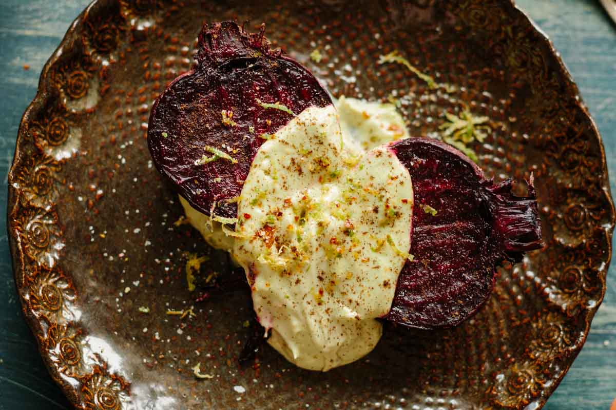 Zoomed-in view of roasted beet halves covered in creamy avocado habanero sauce, sprinkled with lime zest and chile salt on a patterned brown plate.