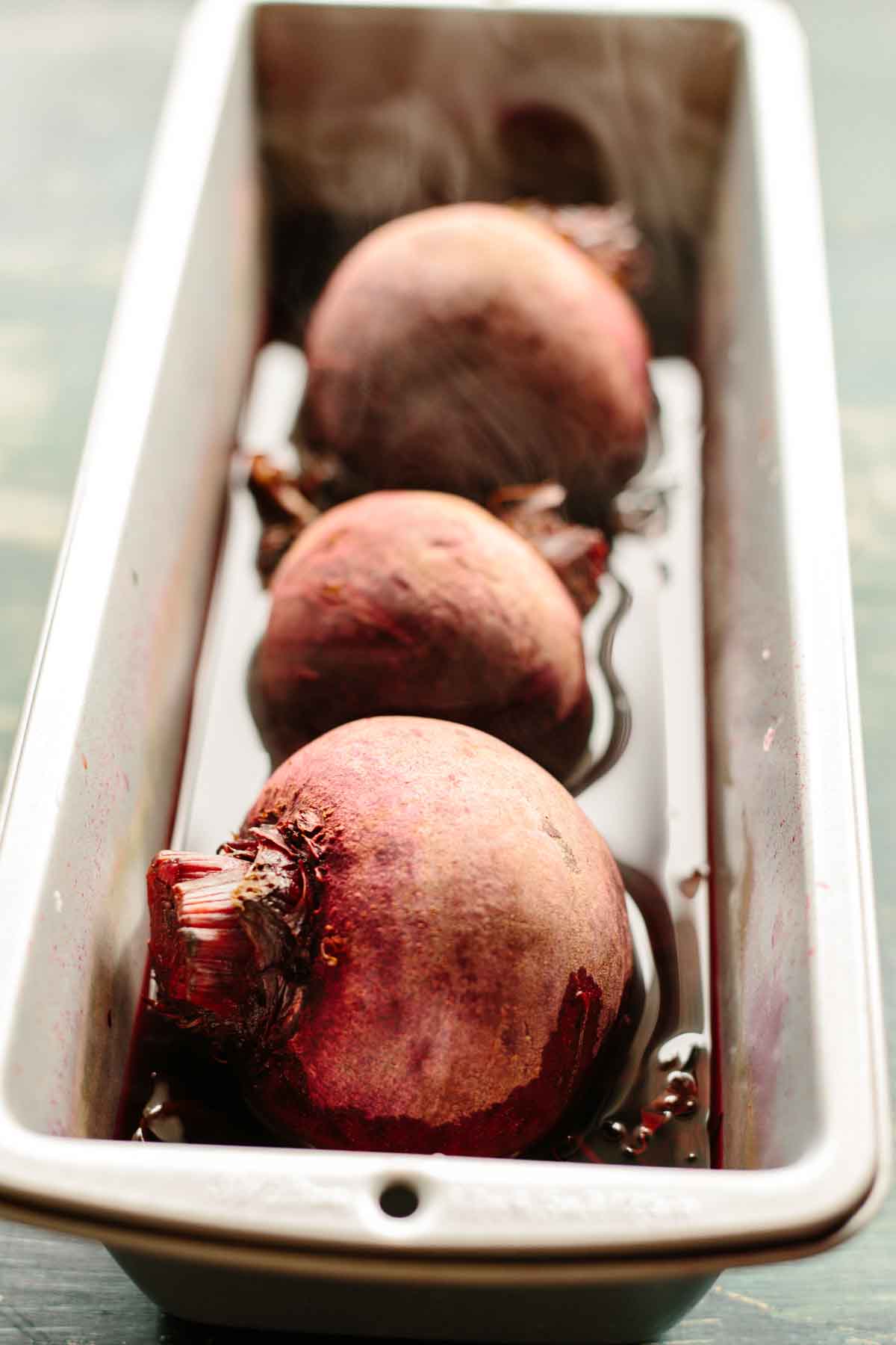Whole raw beets steaming in a loaf pan filled with water, showing the first step of the oven-roasting method.