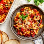 A shallow bowl filled with roasted tomatoes and beans, garnished with fresh basil and grated cheese, served with slices of crusty bread.