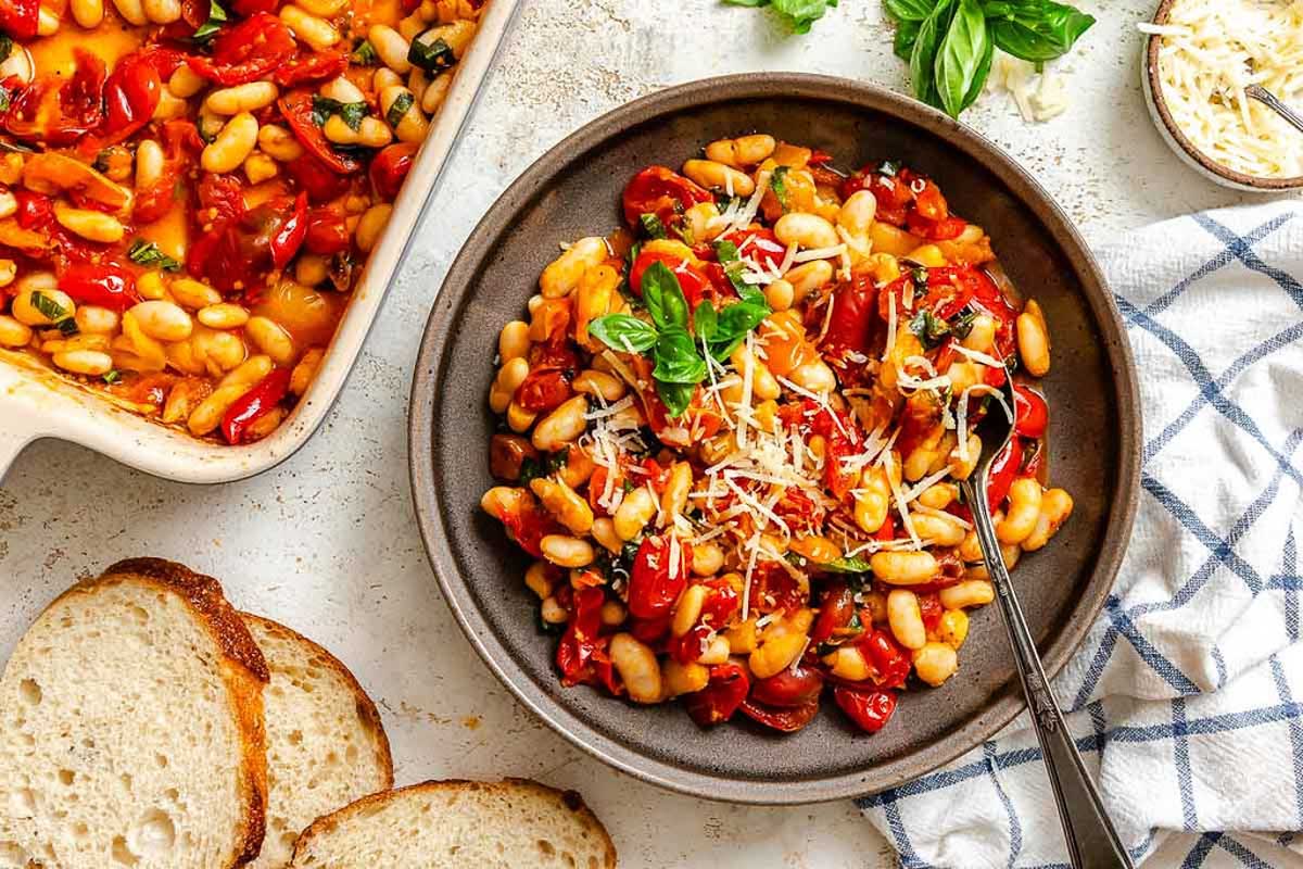 A shallow bowl filled with roasted tomatoes and beans, garnished with fresh basil and grated cheese, served with slices of crusty bread.