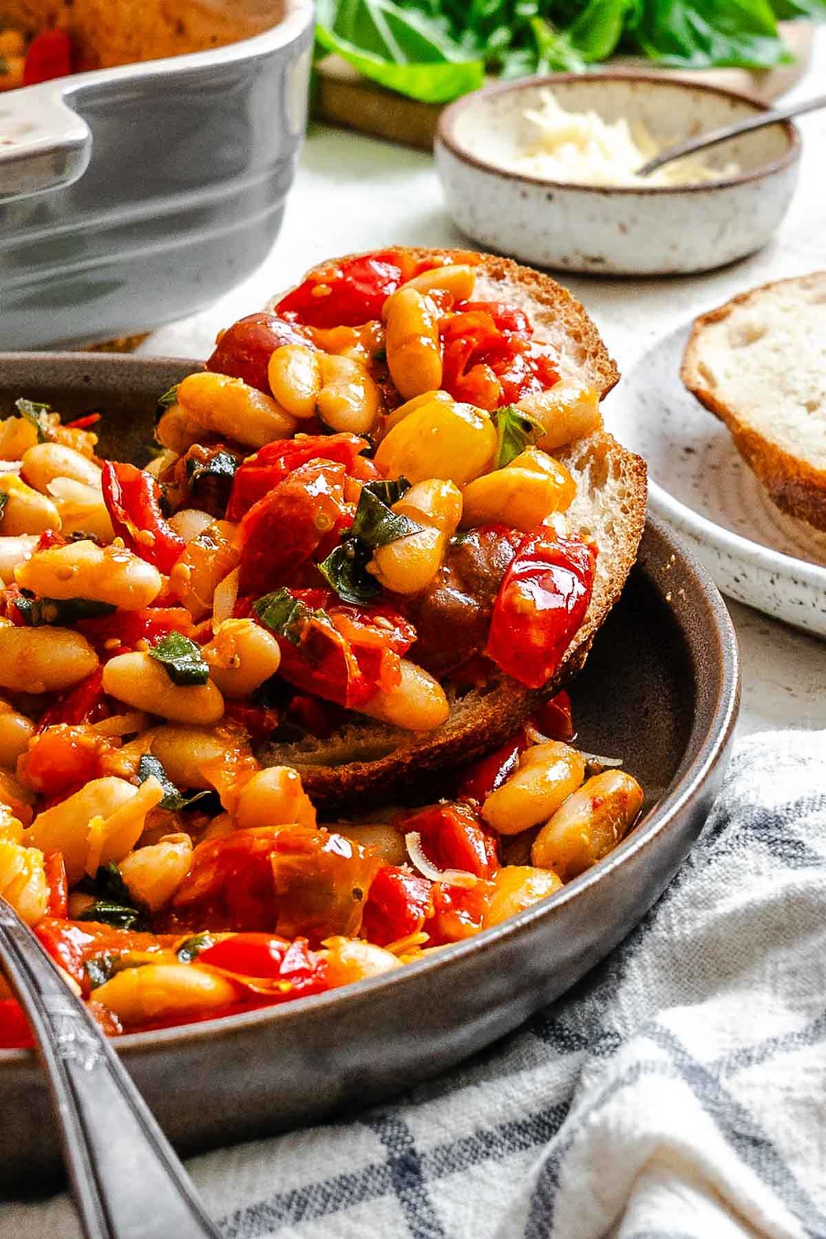 A slice of bread topped with roasted tomatoes and cannellini beans, resting in a bowl with extra sauce and beans around it.