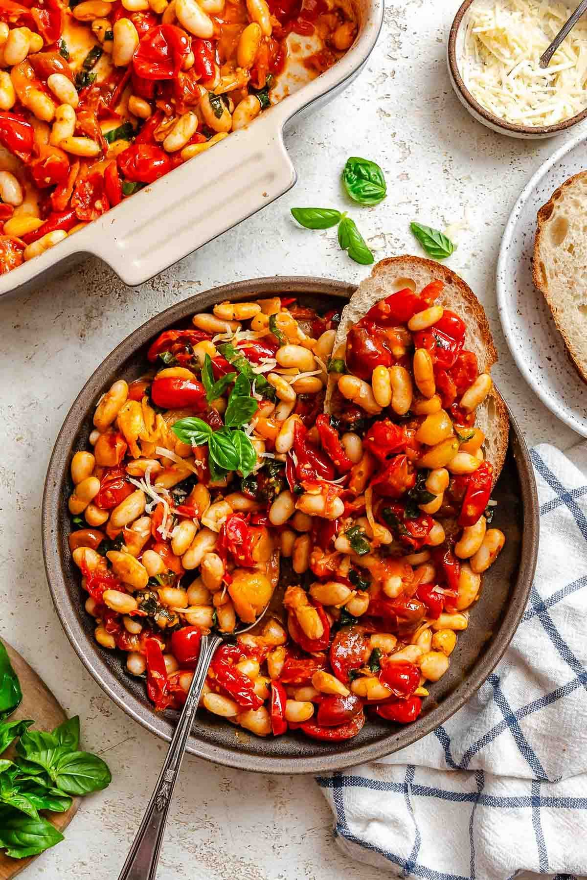Overhead view of roasted tomatoes with beans served in a dark bowl, fresh basil and Parmesan sprinkled on top, with crusty bread on the side.
