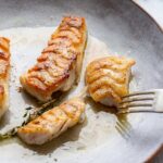 A fork breaking into a flaky, golden brown piece of pan seared tilefish with butter sauce on a gray plate.