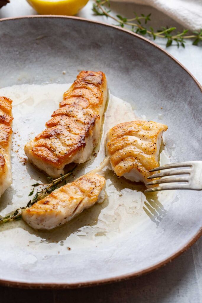 A fork breaking into a flaky, golden brown piece of pan seared tilefish with butter sauce on a gray plate.