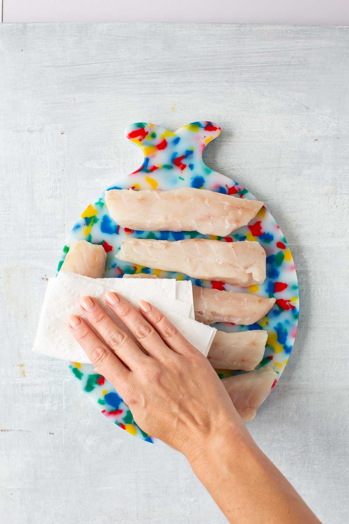 A hand patting raw tilefish fillets dry with paper towels on a colorful fish-shaped cutting board.