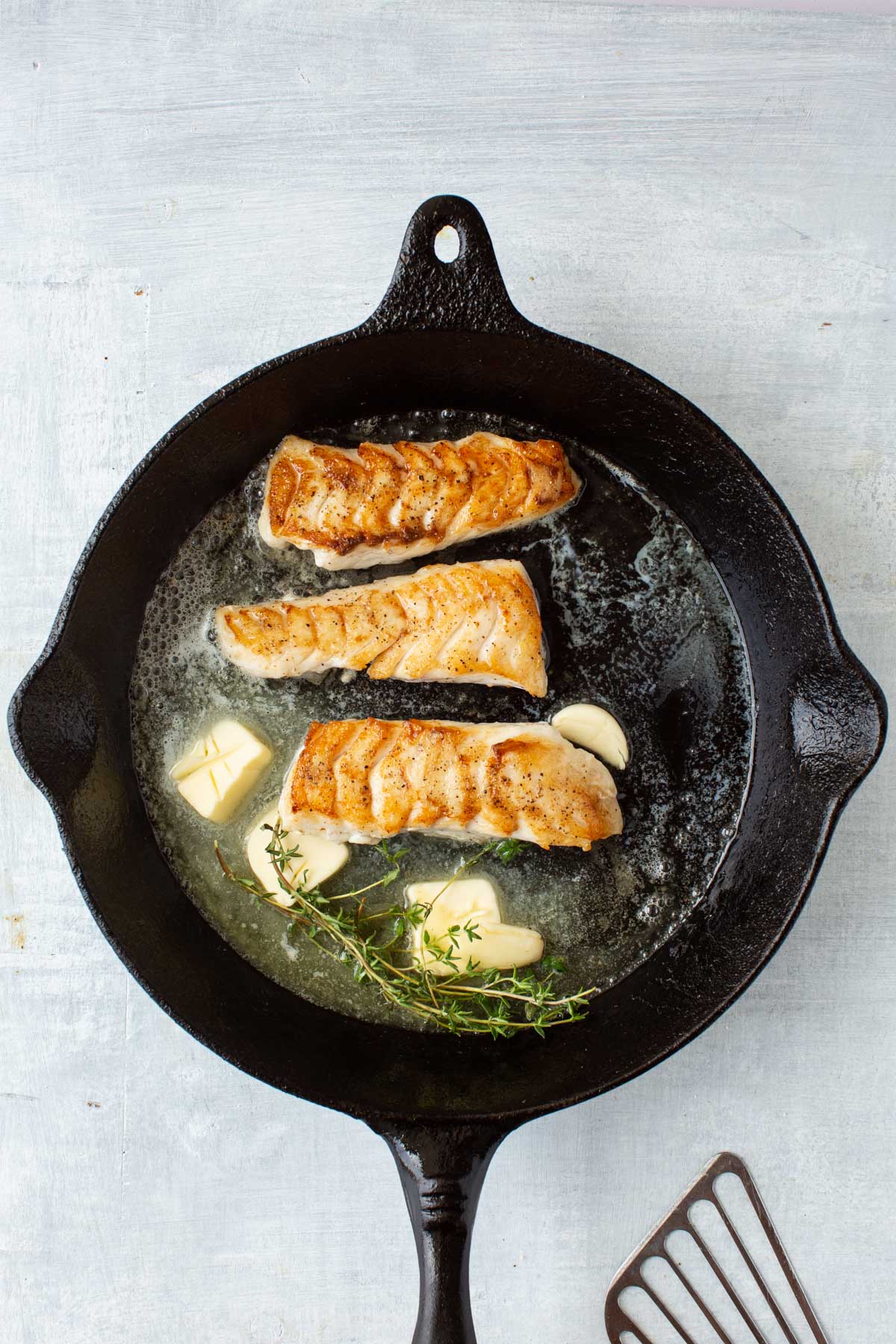 Tilefish fillets browning in a skillet with melted butter, garlic cloves, and fresh thyme sprigs.