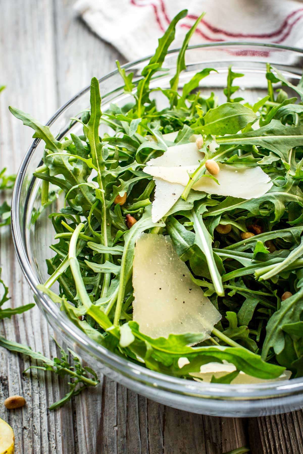 Bright and fresh arugula salad with pine nuts and shaved parmesan in a glass bowl, surrounded by a tea towel and rustic surface.