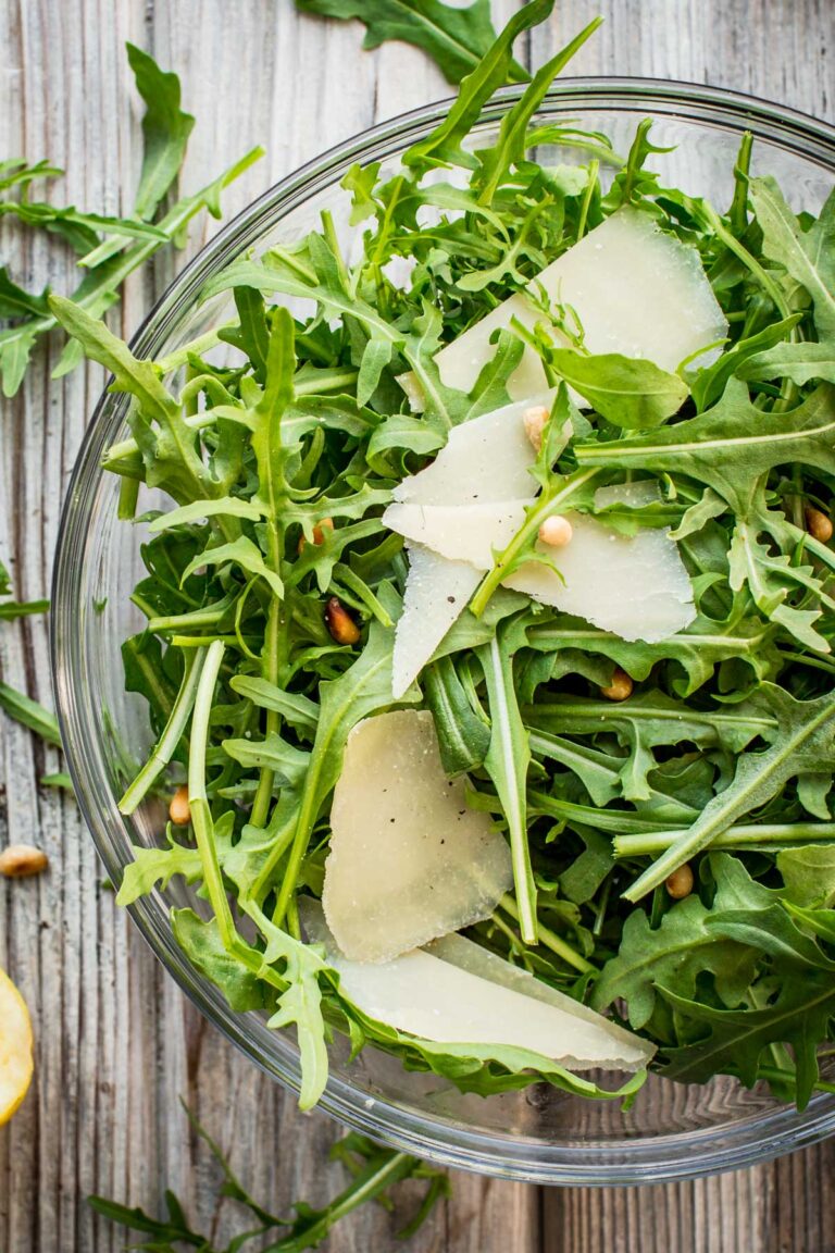 Close-up of arugula leaves, toasted pine nuts, and large shaved parmesan slices in a clear glass bowl.
