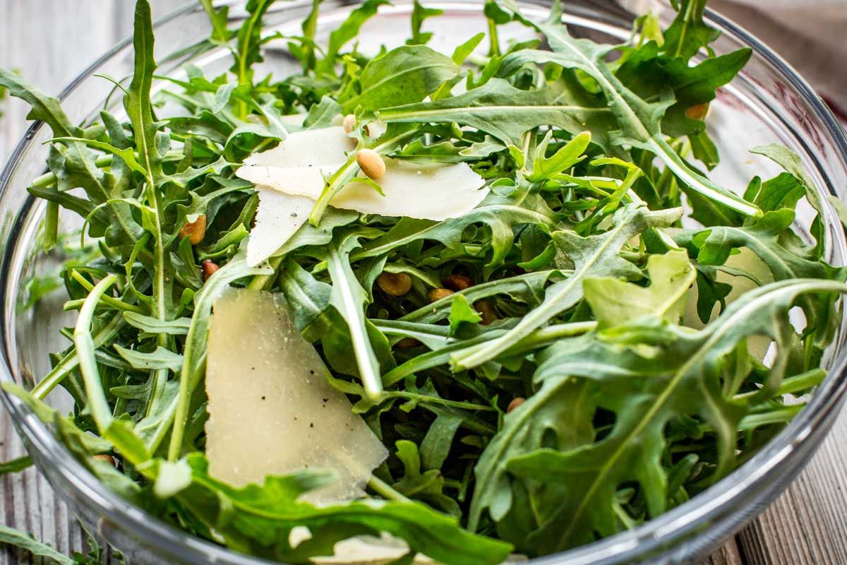 Angled side view of a fresh arugula salad tossed with pine nuts and parmesan in a glass mixing bowl on a wooden table.
