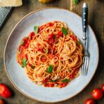 Overhead photo of spaghetti al pomodoro on a gray plate - A serving of spaghetti tossed with fresh tomato sauce and garnished with basil leaves, surrounded by fresh tomatoes, parmesan cheese, and basil on a rustic wooden table.