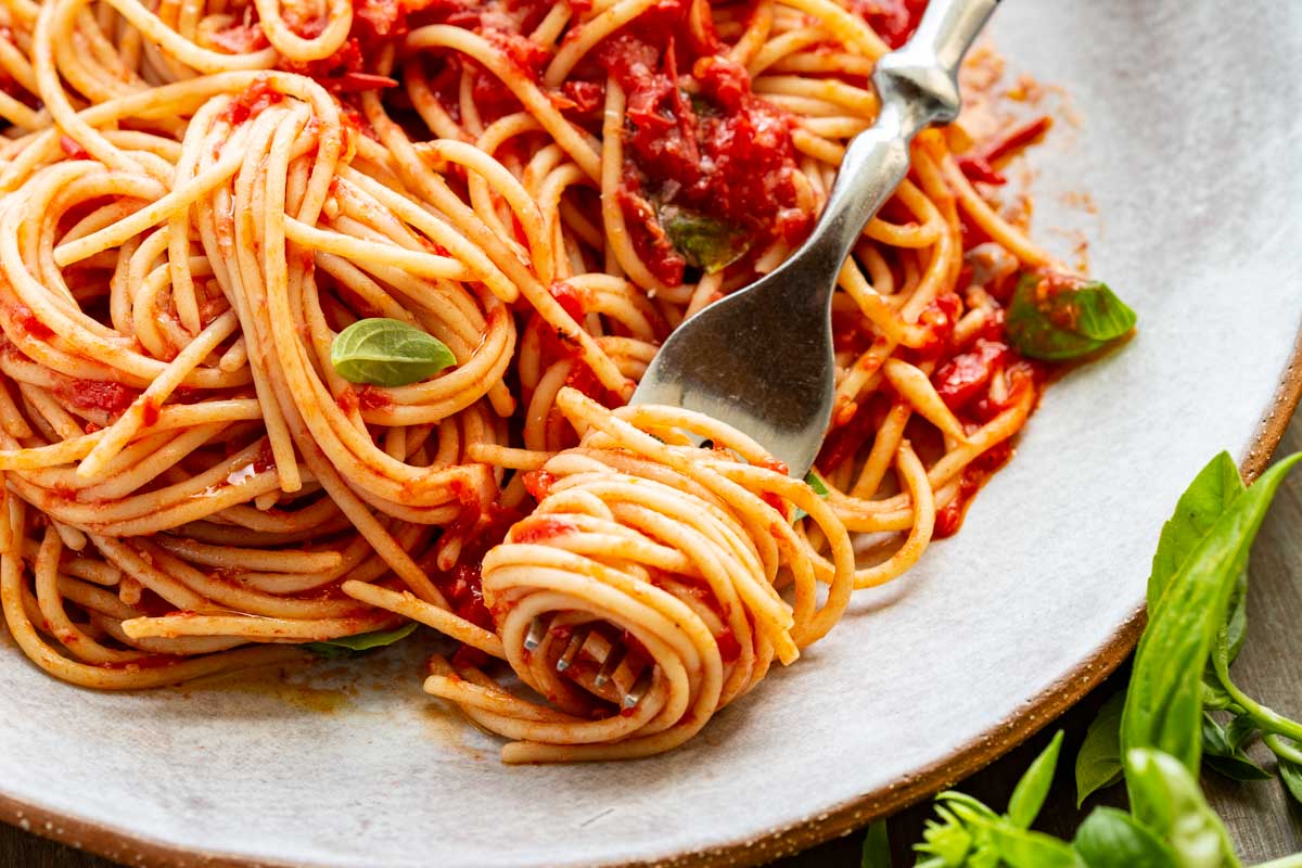 Angled close-up of spaghetti with fork - A fork twirls spaghetti coated in bright red pomodoro sauce, with bits of basil and parmesan visible on a shallow gray plate.