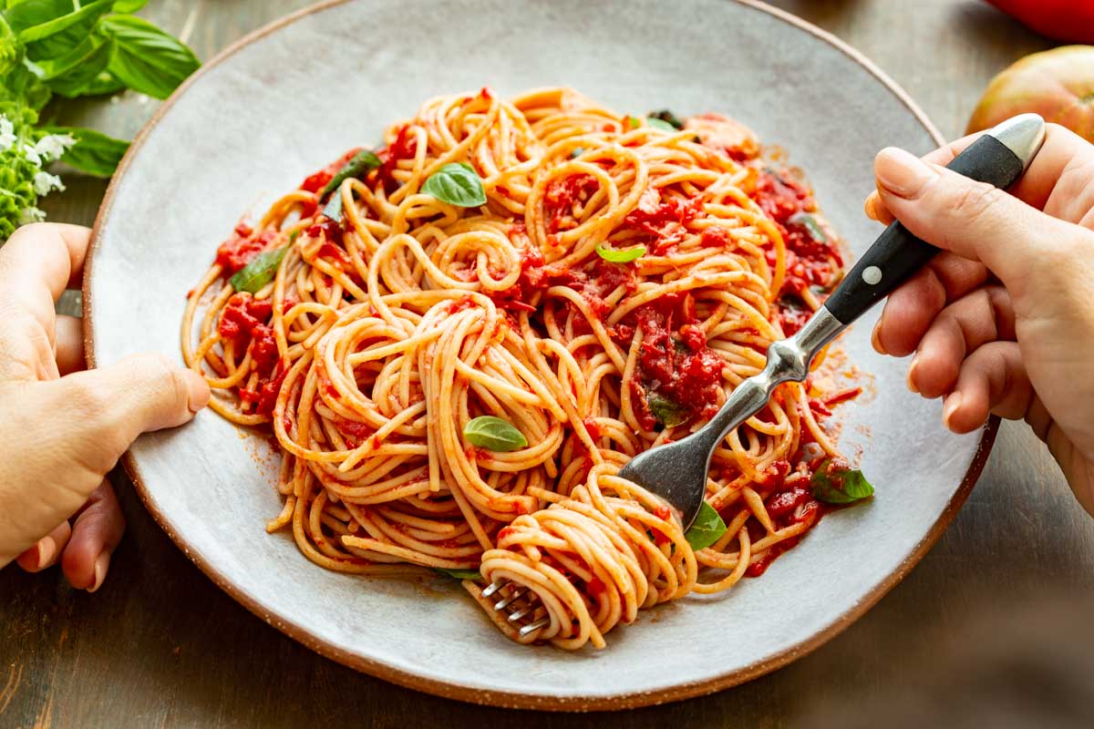 Hands holding a plate of spaghetti al pomodoro – A person twirls spaghetti with a fork while holding a plate full of pasta in fresh tomato sauce with basil leaves.