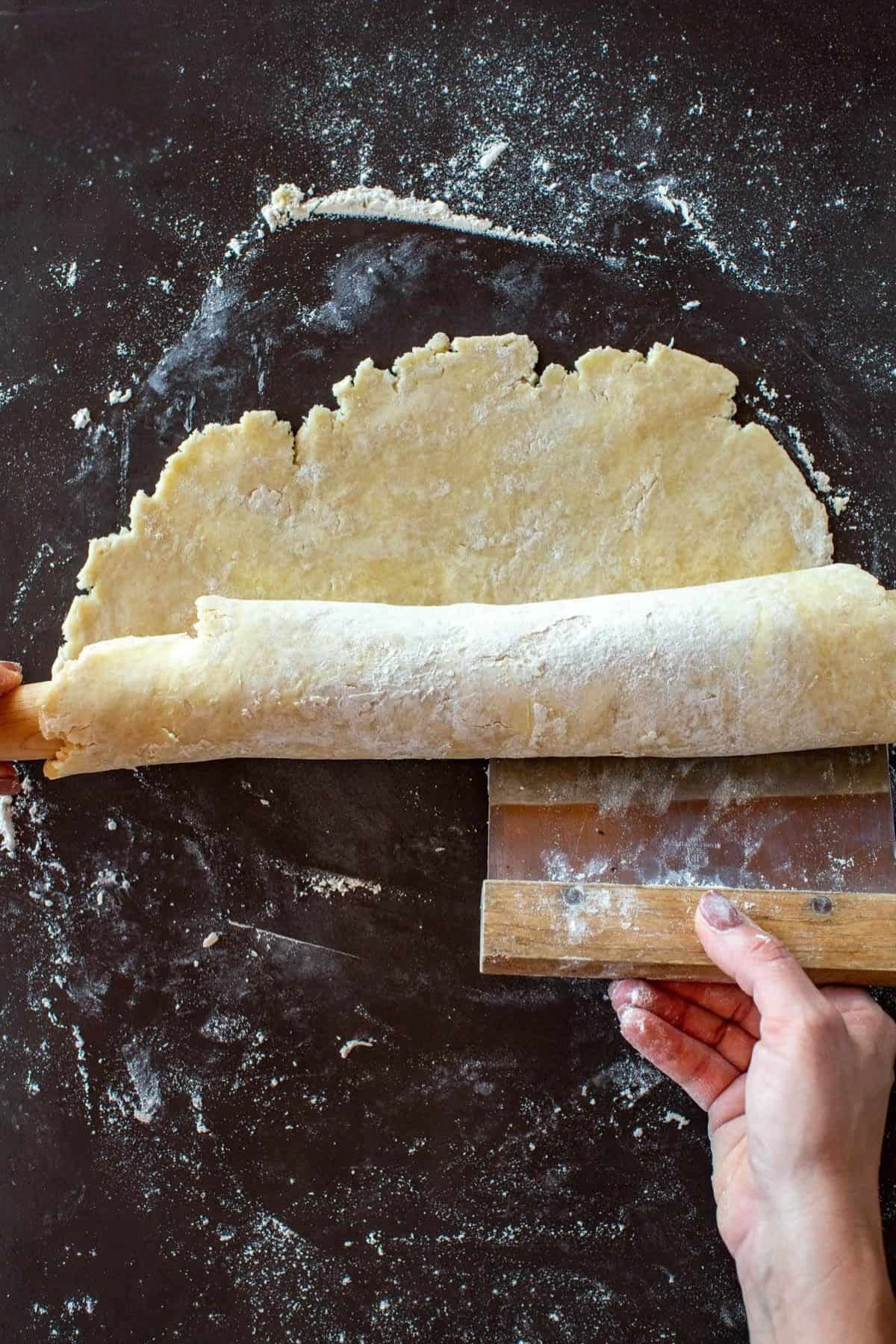 Using a bench scraper to scrape rolled out flaky pie dough off work surface.