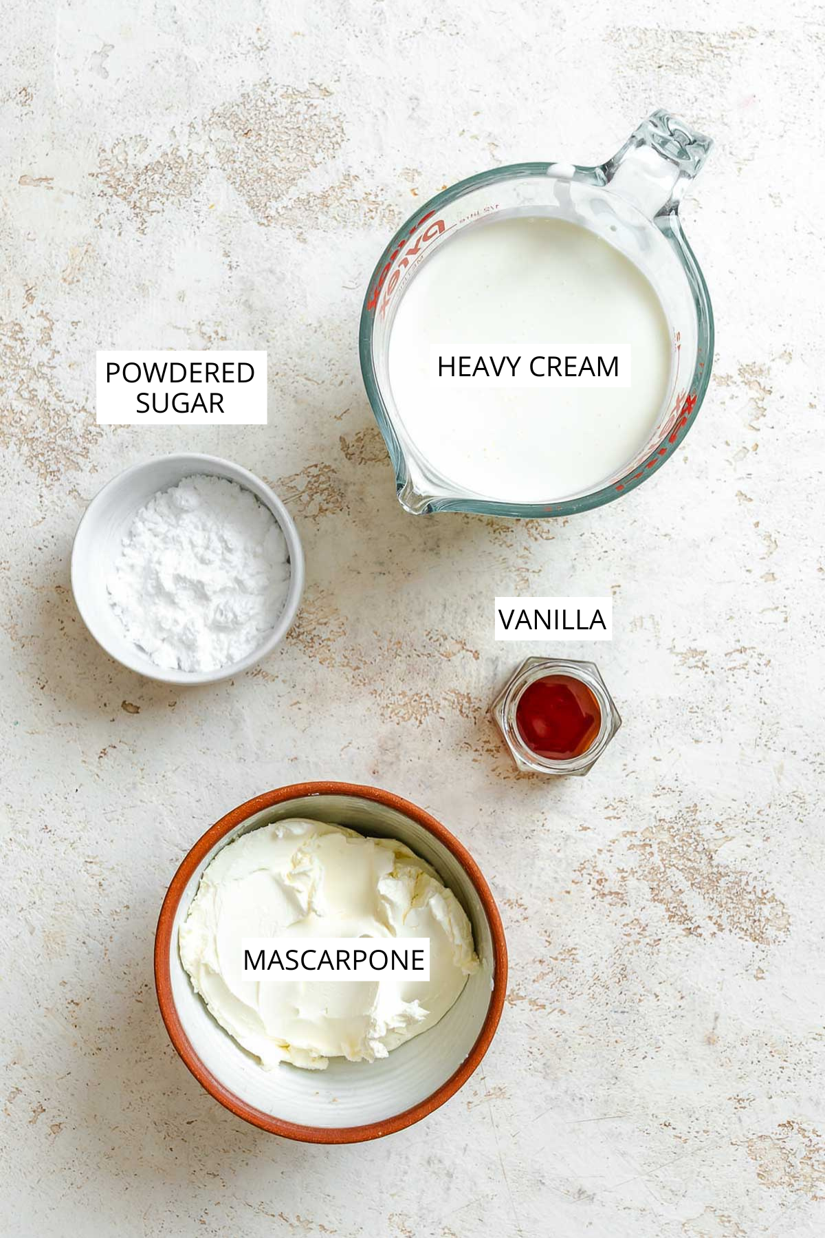 Overhead view of mascarpone, powdered sugar, vanilla, and heavy cream measured out in bowls and a glass measuring cup on a light surface.
