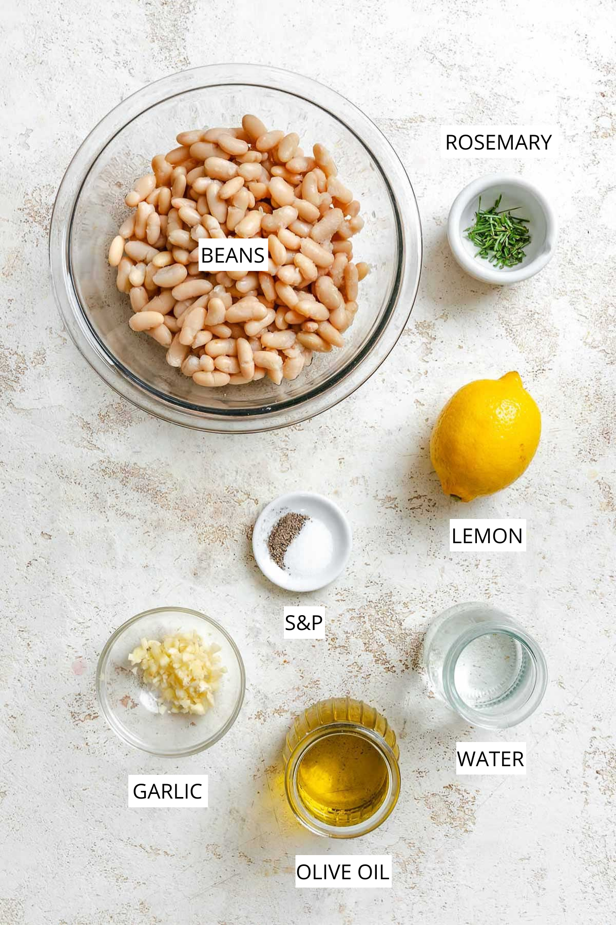Ingredients for the dip laid out on a light surface: a bowl of cannellini beans, fresh rosemary, lemon, minced garlic, olive oil, salt, pepper, and water.