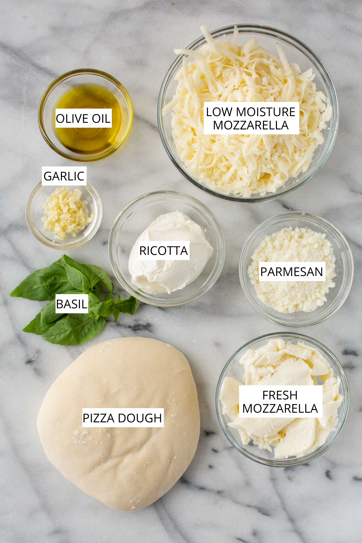 Ingredients overhead - A marble surface with labeled ingredients for pizza bianca, including a dough ball, fresh mozzarella, shredded mozzarella, ricotta, parmesan, garlic, olive oil, and basil.