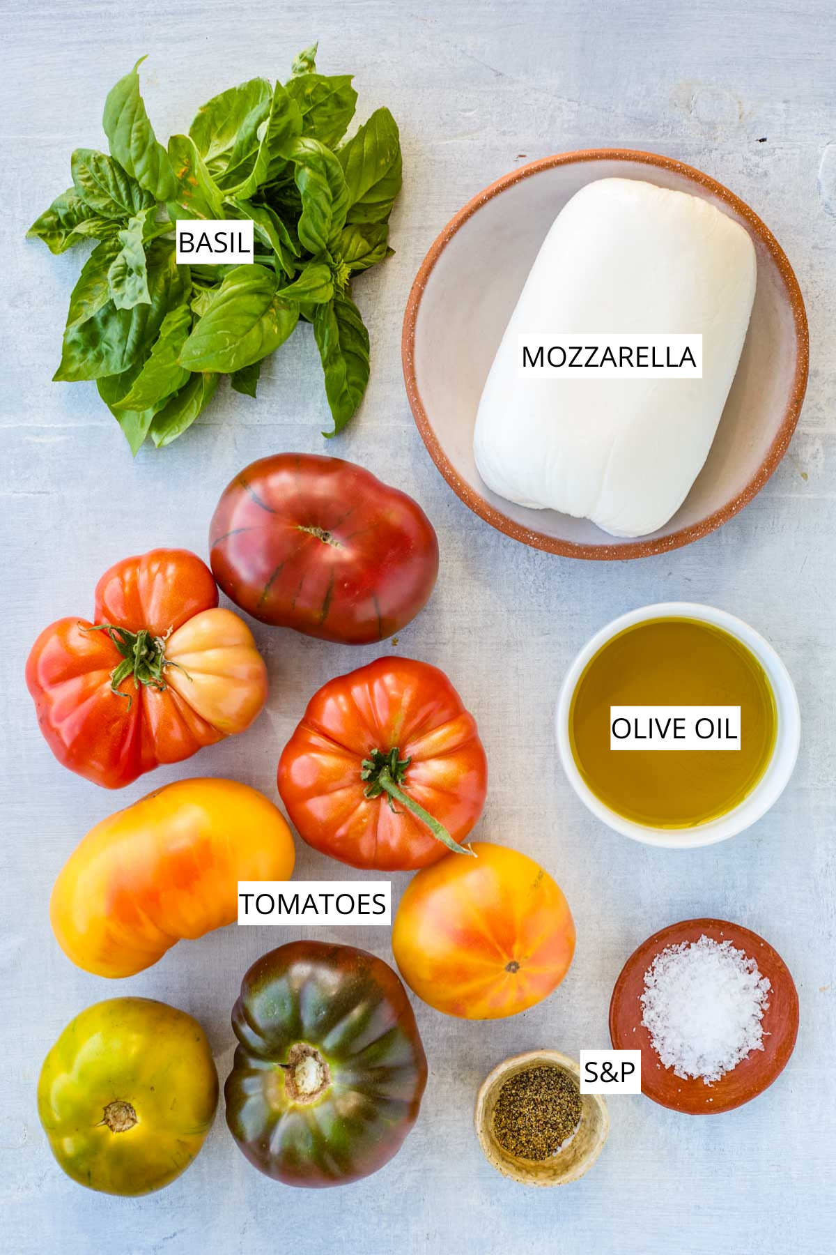 Ingredients for Caprese salad laid out, including heirloom tomatoes, fresh mozzarella, basil, olive oil, salt, and pepper with labels.