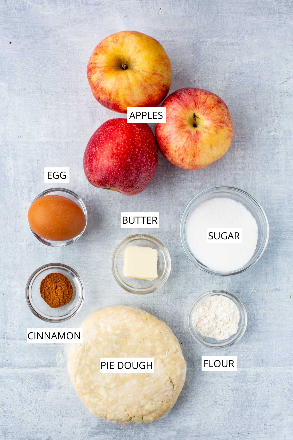 Overhead of the labeled ingredients for apple crostata, including apples, pie dough, butter, flour, sugar, cinnamon, and an egg.