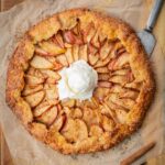 Freshly baked apple crostata on parchment paper with a scoop of vanilla ice cream melting in the center, surrounded by cinnamon sticks and a whole apple on a wooden board.