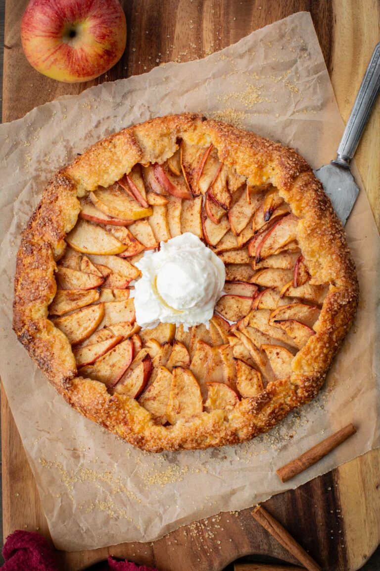 Freshly baked apple crostata on parchment paper with a scoop of vanilla ice cream melting in the center, surrounded by cinnamon sticks and a whole apple on a wooden board.