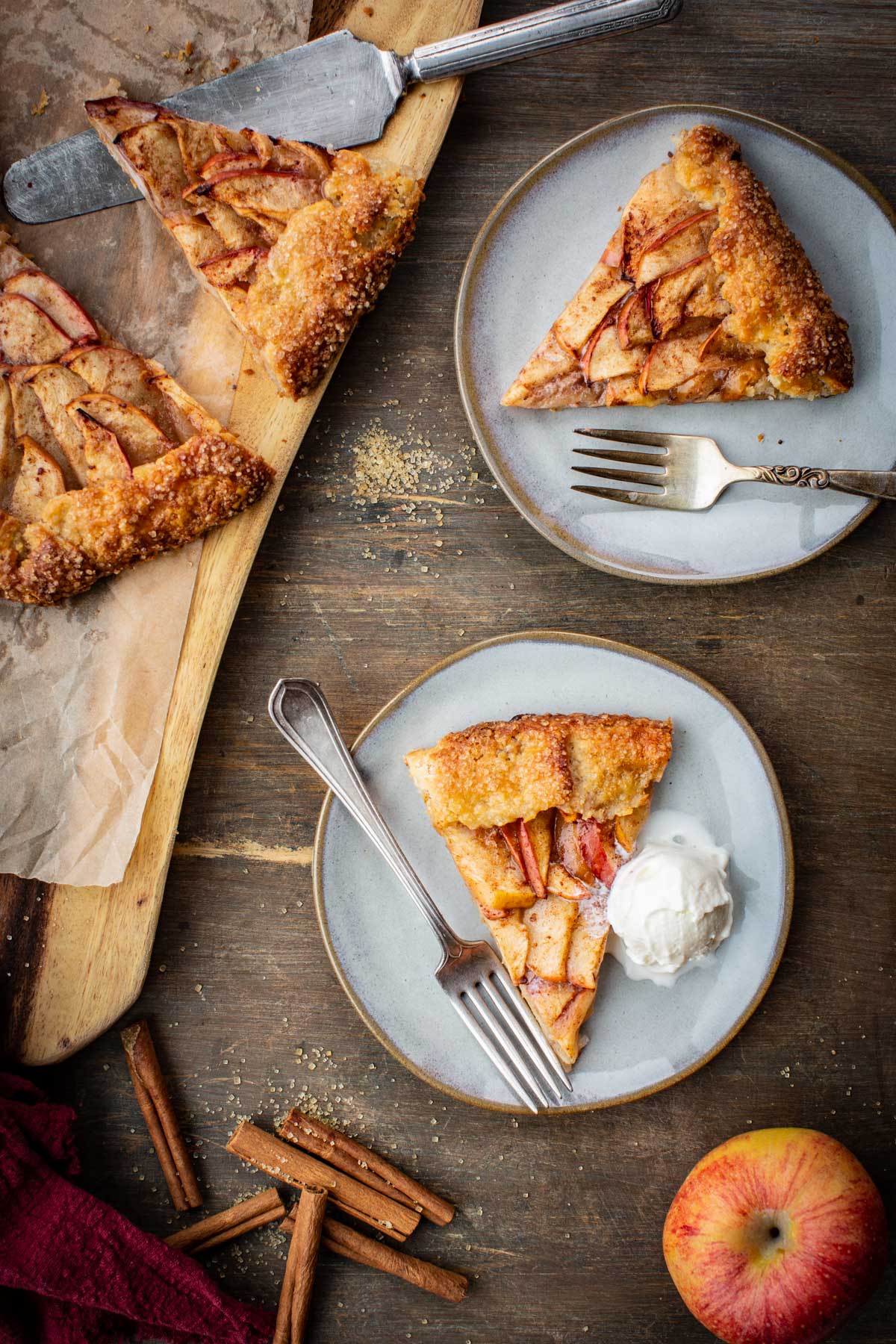 Two plated slices of apple crostata on gray plates, one topped with vanilla ice cream, with a pie server and cinnamon sticks scattered nearby.