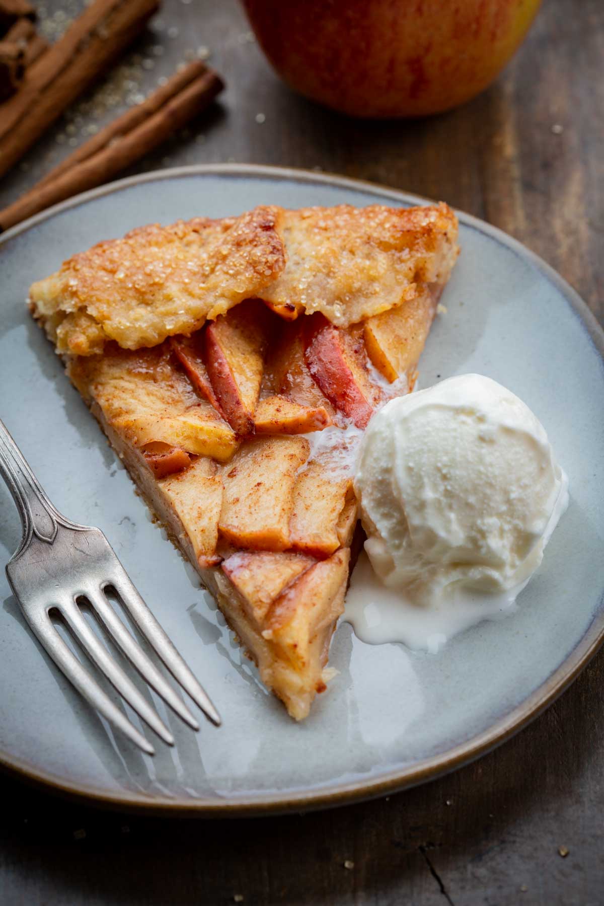 A close-up of a single slice of apple crostata on a gray plate, served with a scoop of melting vanilla ice cream and a fork.