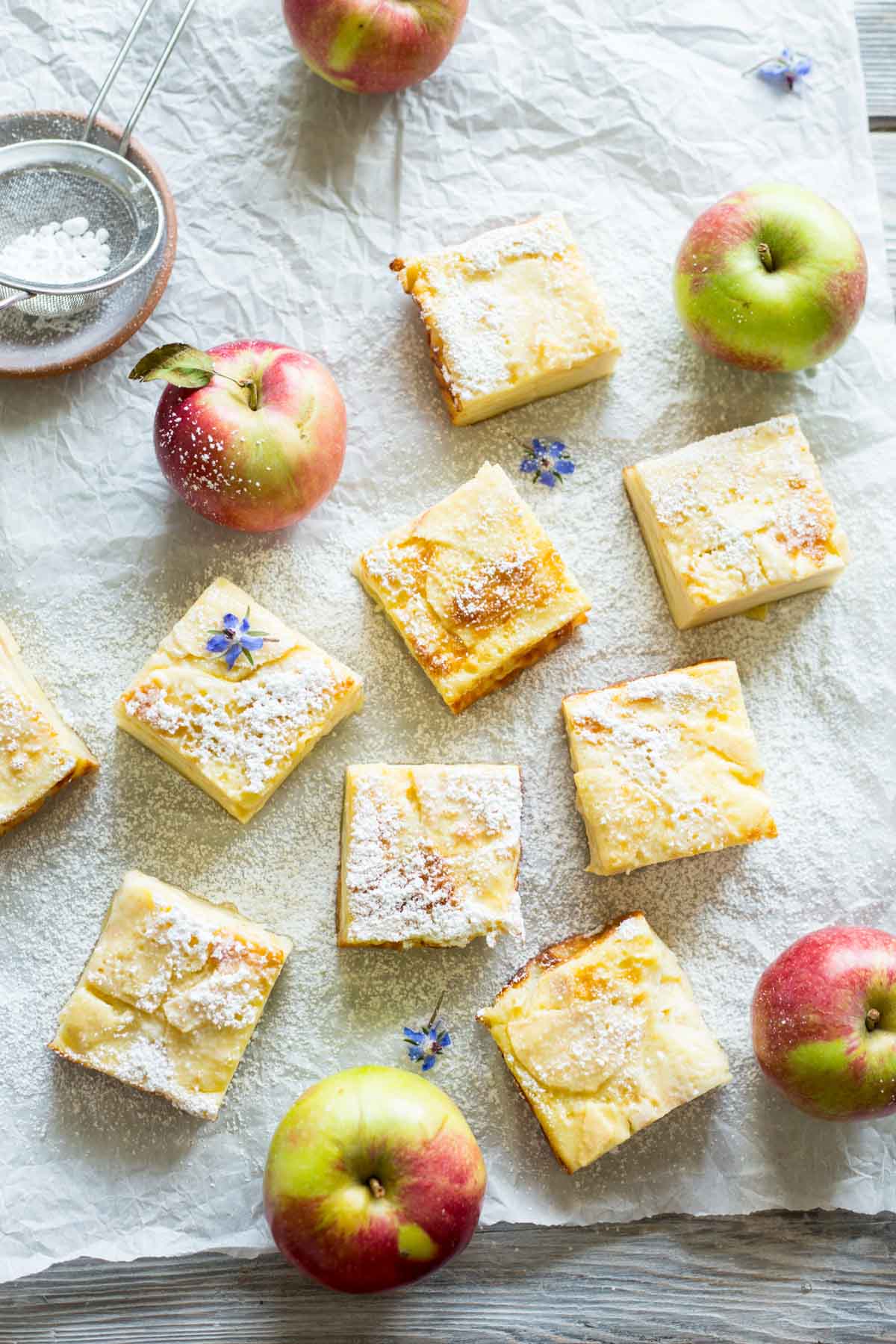 Squares of apple custard bars dusted with powdered sugar on crinkled parchment, surrounded by fresh apples and a small sieve.