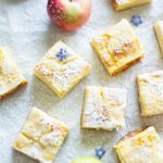 Close-up view of apple custard bars topped with powdered sugar and small edible flowers, with apples scattered around.