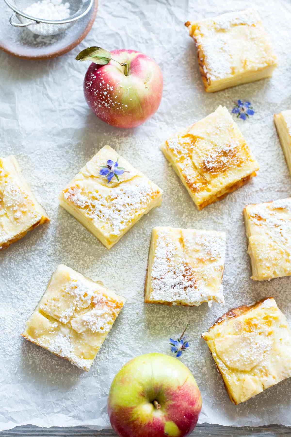 Close-up view of apple custard bars topped with powdered sugar and small edible flowers, with apples scattered around.