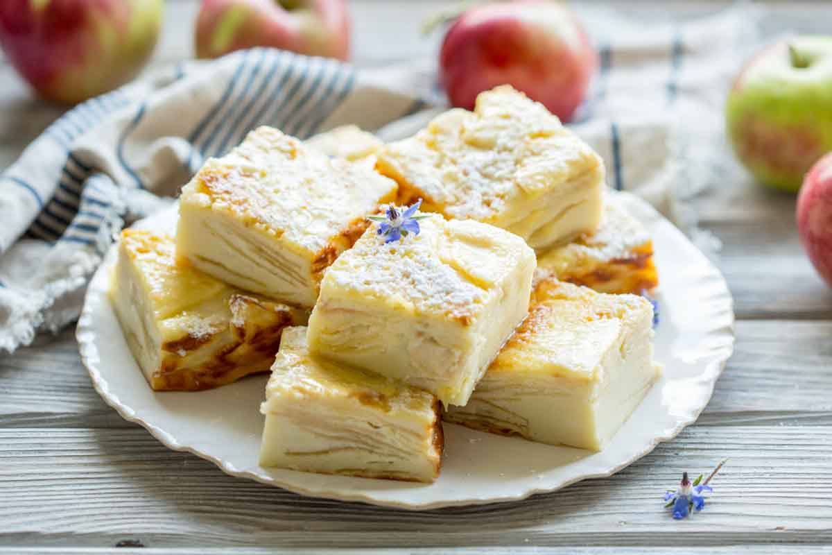 A white plate stacked with thick apple custard bars, dusted with powdered sugar and decorated with a small purple flower.