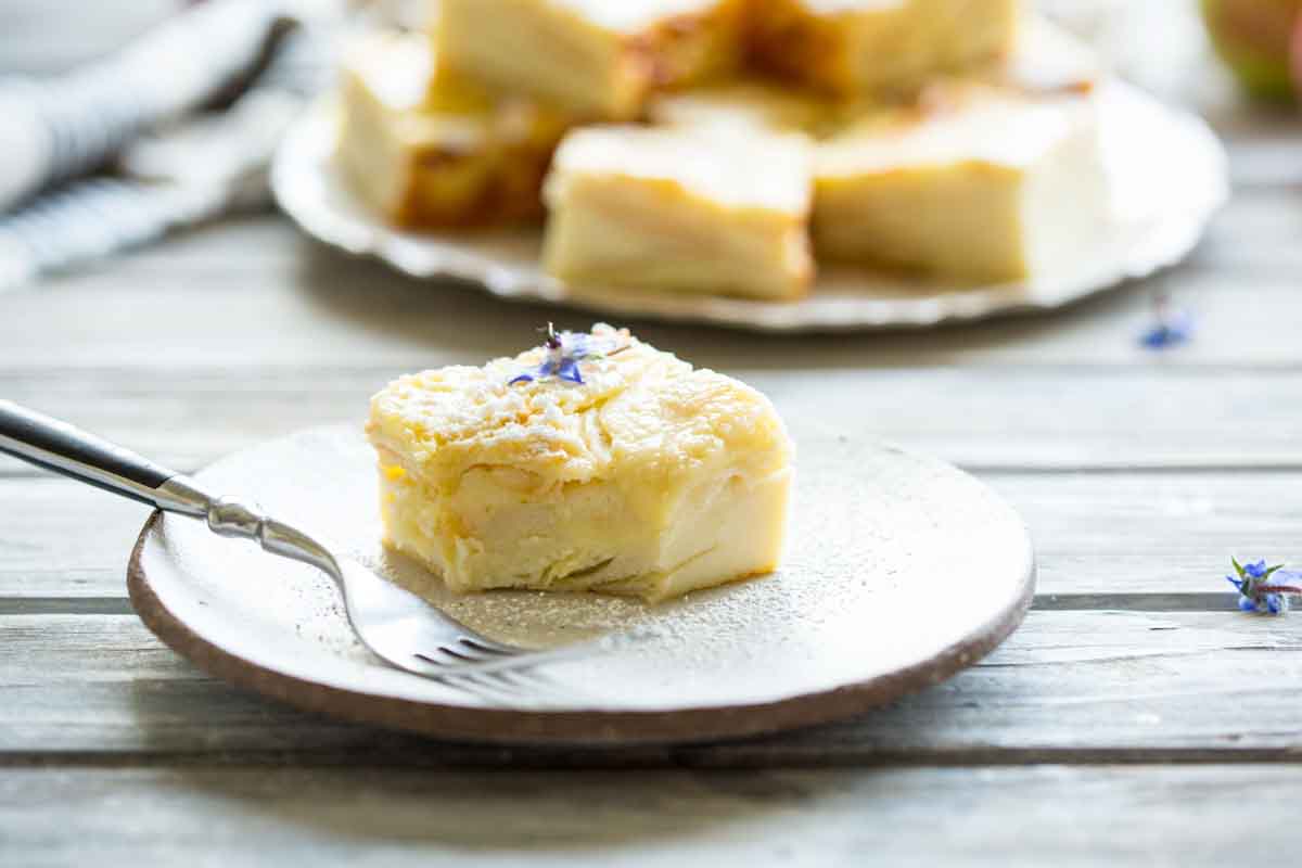 A single apple custard bar on a small plate with a fork, lightly dusted with powdered sugar, with more bars blurred in the background.