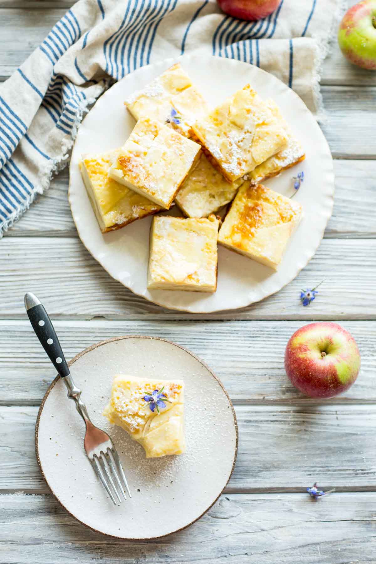 Overhead shot of a serving plate piled with apple custard bars and one plated piece with a fork, styled with apples and a striped cloth.