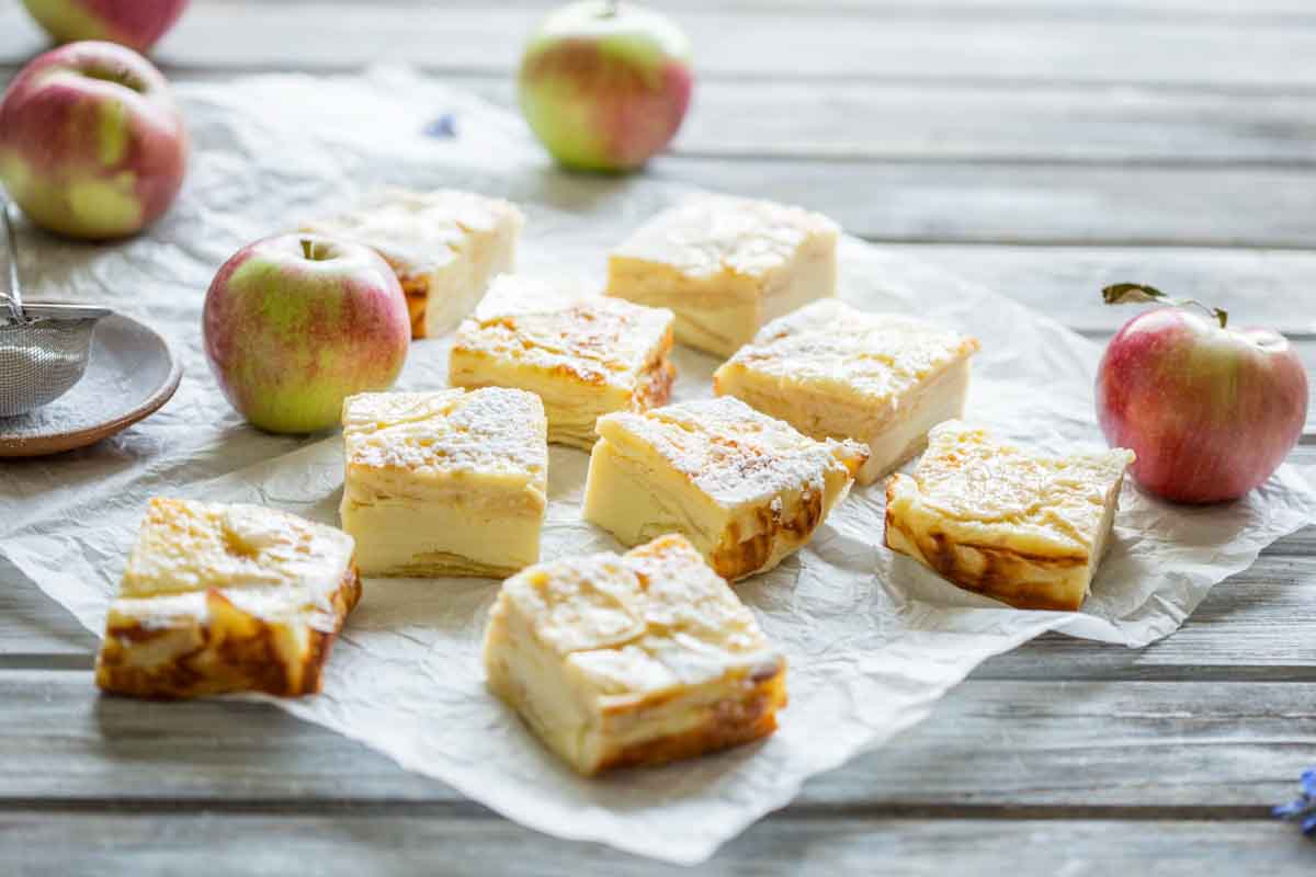 Apple custard bars arranged on parchment paper with whole apples around, showing the golden tops and creamy layered interiors.