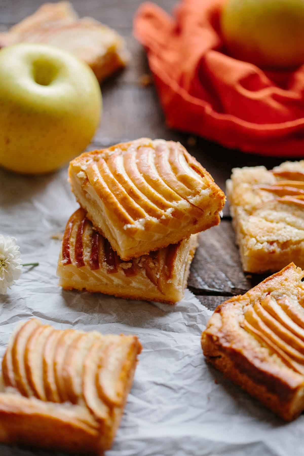 Two apple almond shortbread bars stacked on parchment paper, surrounded by whole apples and orange linen, highlighting the crisp edges and tender apple topping.