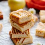 Stacked apple almond shortbread bars on parchment paper with whole apples in the background, showcasing the golden brown crust and sweet apple filling.