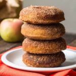 A tall stack of four baked apple cider donuts coated in cinnamon sugar on a white plate with a red napkin, fresh apples in the background.
