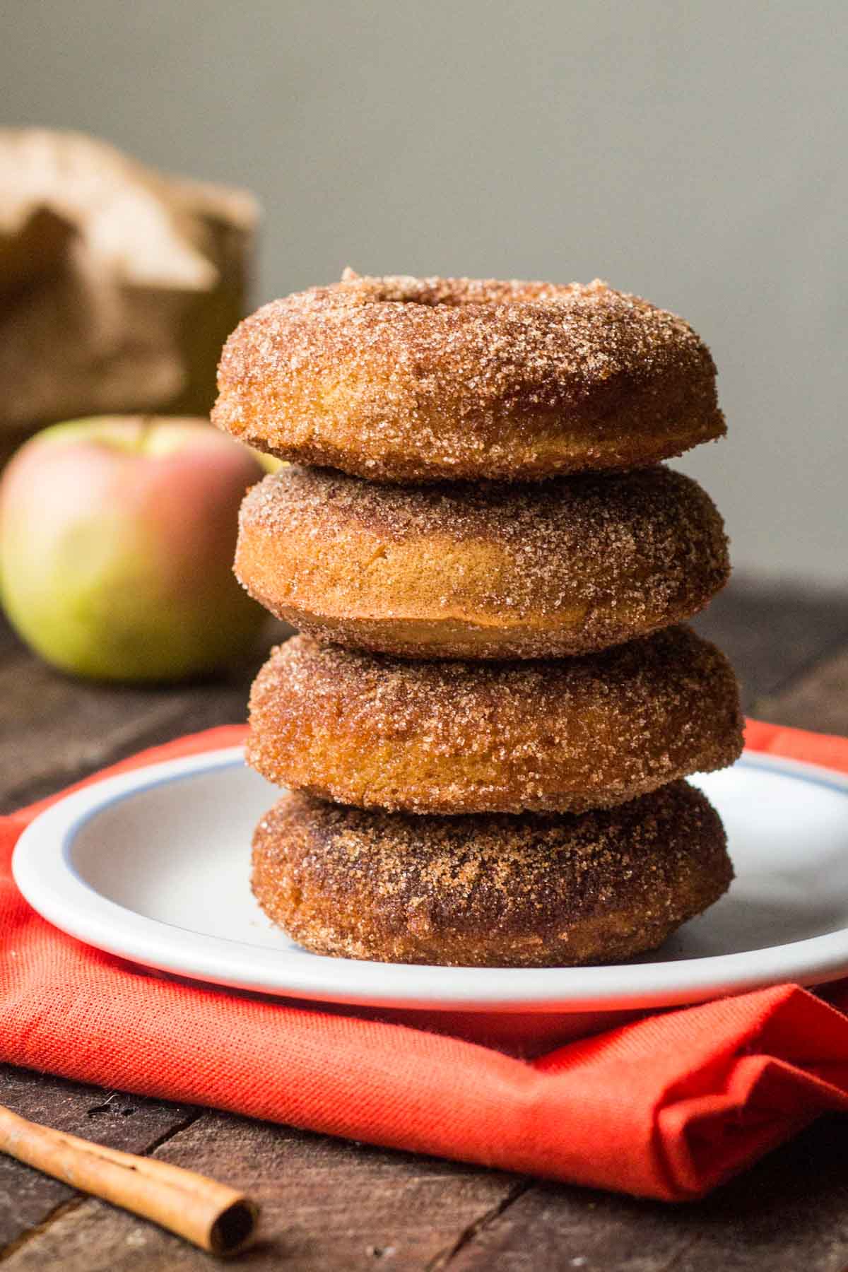 A tall stack of four baked apple cider donuts coated in cinnamon sugar on a white plate with a red napkin, fresh apples in the background.