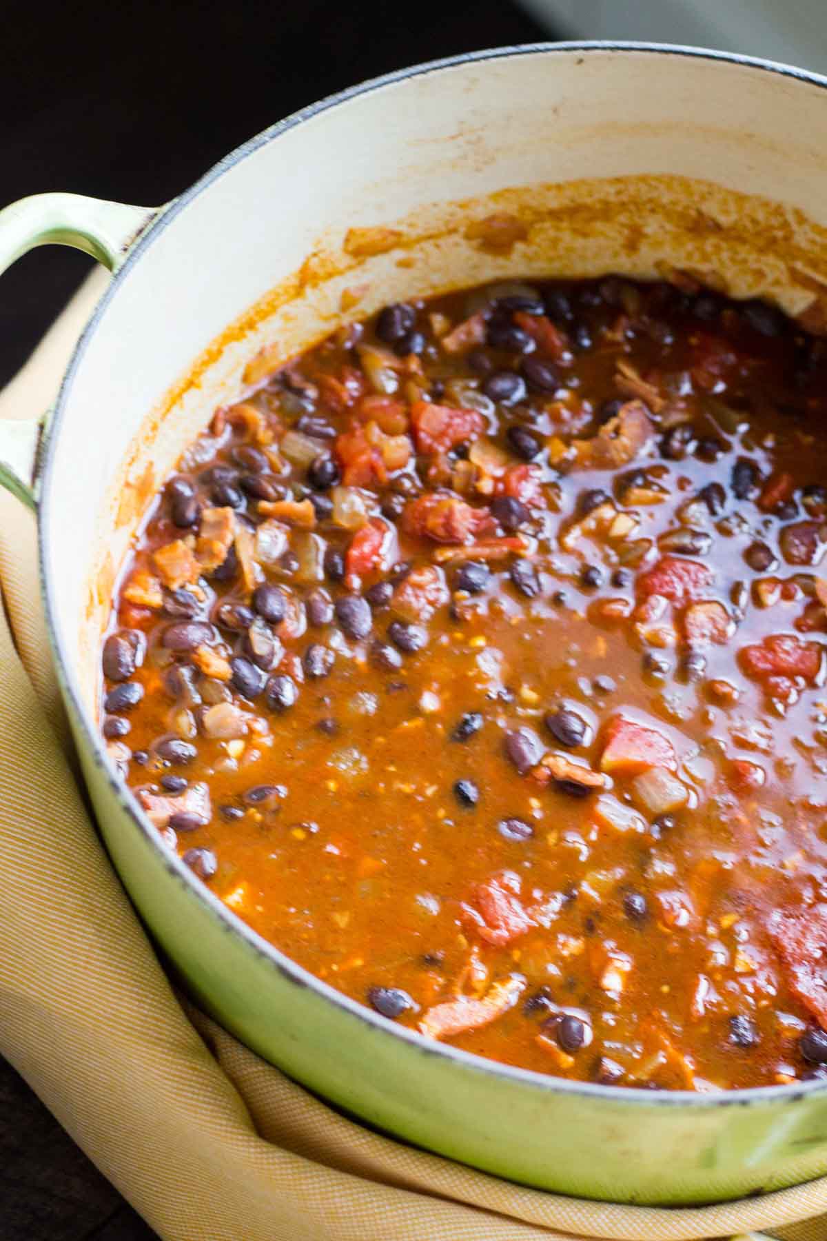 A large Dutch oven filled with simmering black bean soup with tomatoes, onions, and bacon in a rich broth.