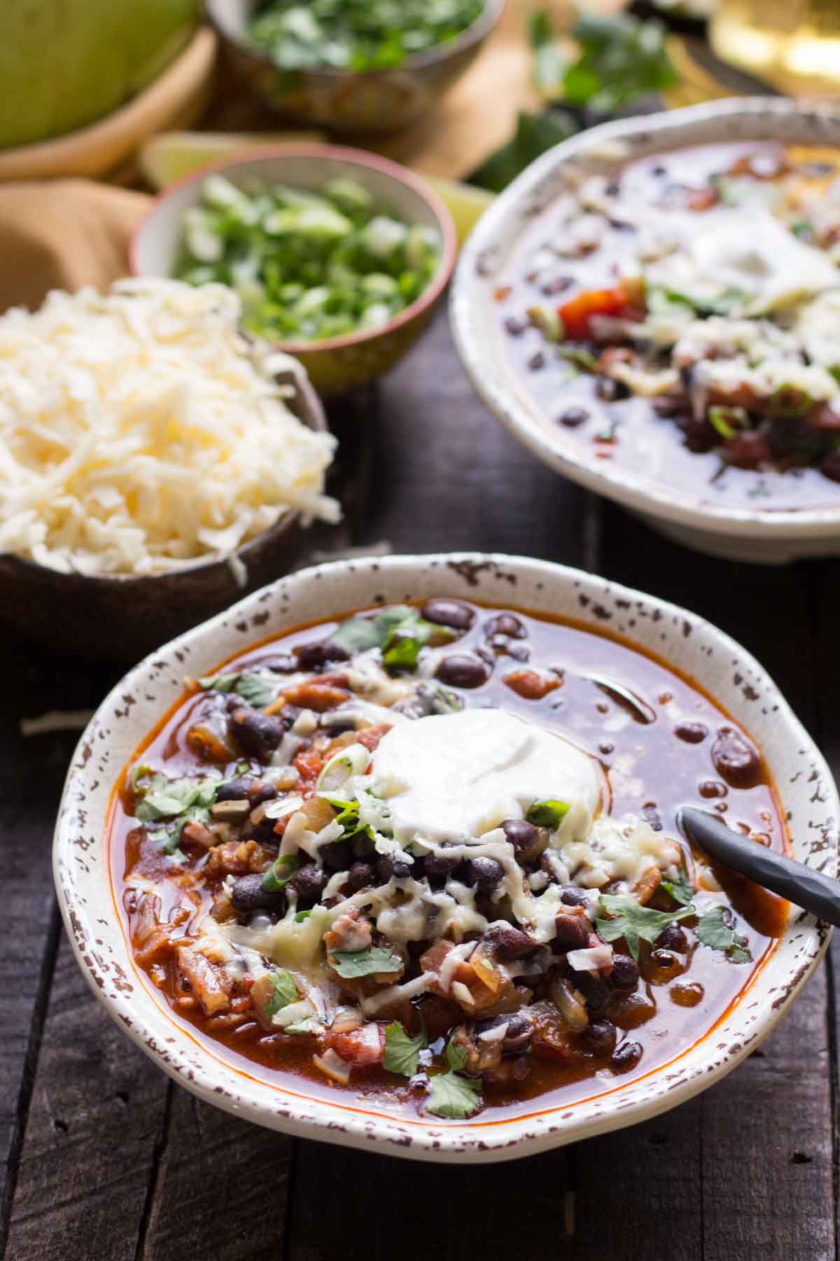 A bowl of black bean soup topped with sour cream, shredded cheese, and fresh cilantro, with bowls of shredded cheese and green onions in the background.