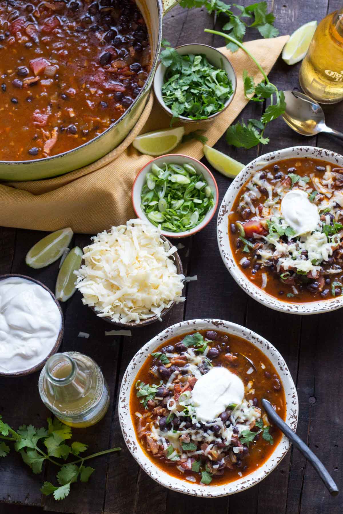 Overhead view of a pot of black bean soup with two bowls served alongside toppings of cilantro, green onions, shredded cheese, lime wedges, and sour cream.