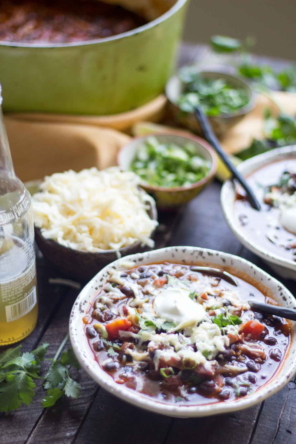 Two bowls of black bean soup on a rustic table, surrounded by toppings like shredded cheese, cilantro, lime wedges, and sour cream, with the pot of soup in the background.